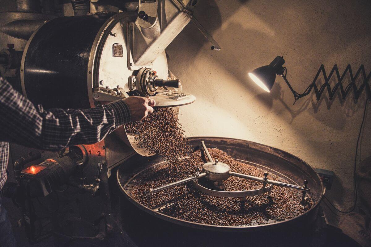 The image captures a moment in the coffee roasting process. A person, visible in side profile, is pouring coffee beans into a roasting machine. The scene is dominated by shades of brown and beige, evoking the warm, inviting aroma of coffee.